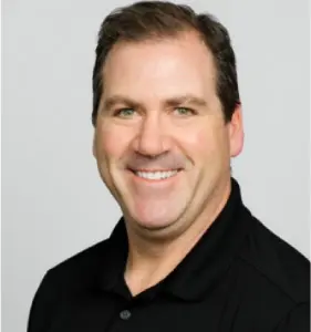 Smiling man with short dark hair wearing a black collared shirt against a light gray background.