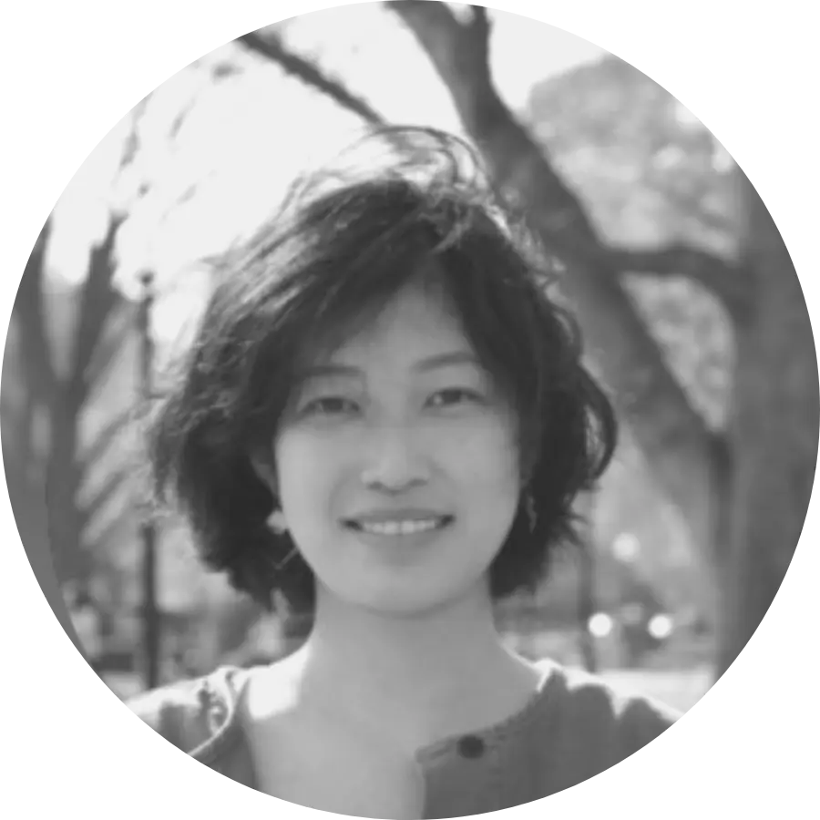 A grayscale portrait of a person with short, tousled hair smiling softly at the camera. They are standing outdoors, with blurred trees and park elements in the background.