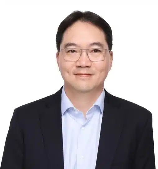 A man with glasses, wearing a dark suit and light blue shirt, smiles at the camera against a plain white background.