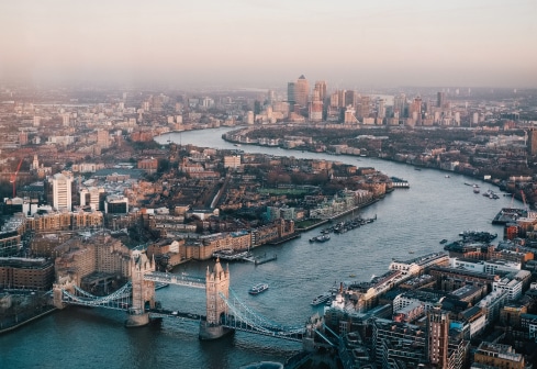 Aerial view of London, featuring the River Thames winding through the city. The iconic Tower Bridge is visible in the foreground, with numerous buildings and the skyline stretching into the distance under a hazy sky.