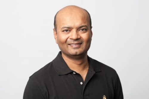 A person with a shaved head and a slight smile is wearing a black collared shirt against a plain white background.