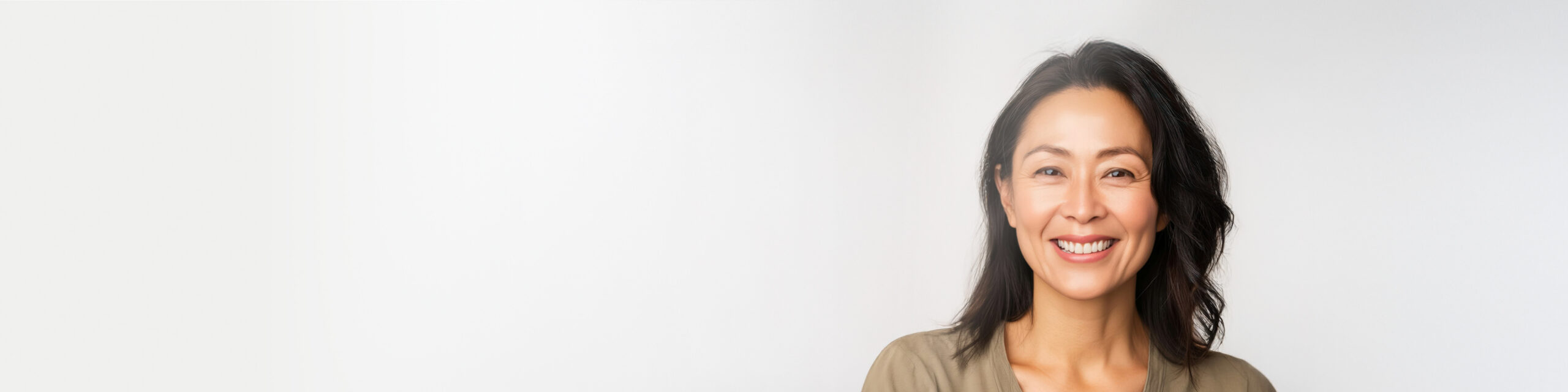 Smiling woman with long black hair stands against a light gray background, wearing a beige top. The image has a soft, minimalistic feel with space on the left side.