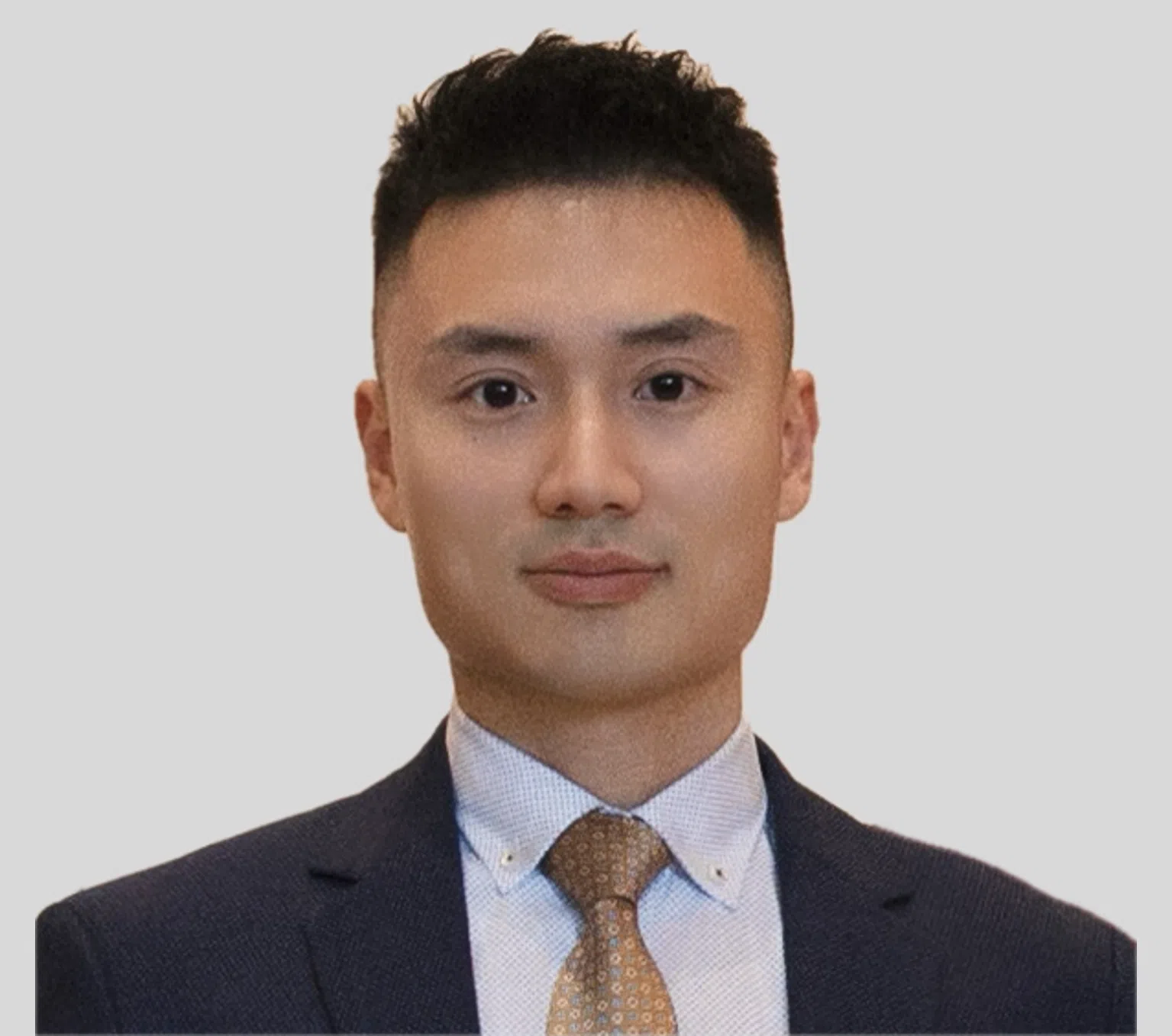 A man with short black hair wearing a dark suit, white dress shirt, and patterned tan tie, posing against a plain light gray background.