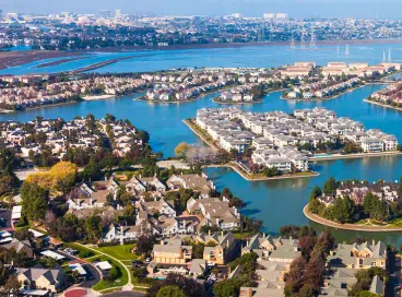 Aerial view of a suburban neighborhood with houses, tree-lined streets, and waterways, set against a backdrop of a large city and a body of water in the distance.