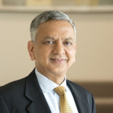 A middle-aged man with short gray hair, wearing a dark suit, light blue shirt, and yellow patterned tie, smiling slightly while looking at the camera, in a softly lit indoor setting.