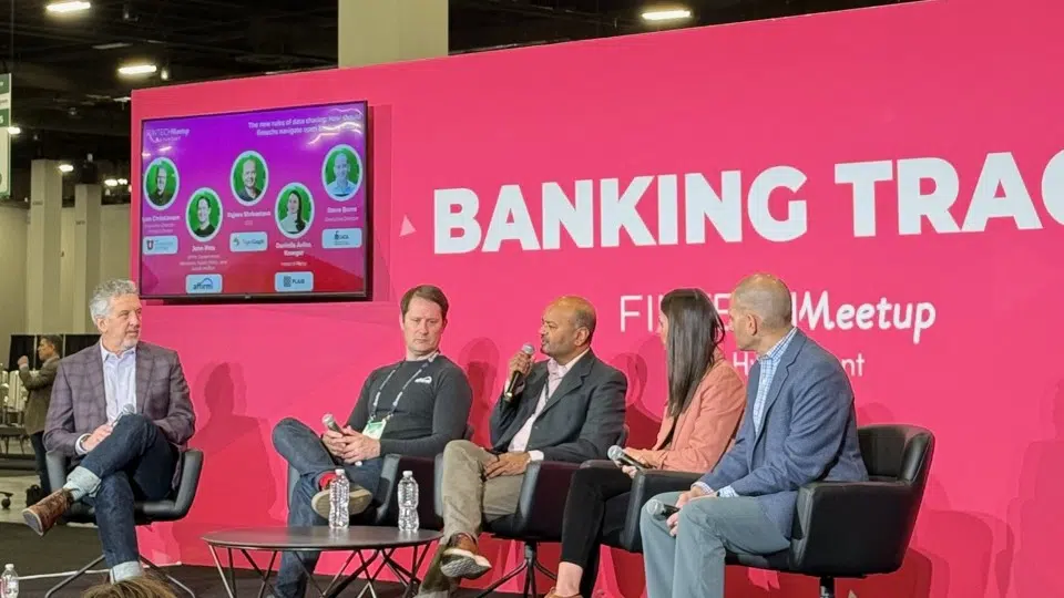 Five people sit on a panel discussing how open banking compliance can be a competitive advantage, on a stage with a bright pink BANKING TRACK backdrop. A screen behind them displays the panel’s topic along with speakers’ names and photos.